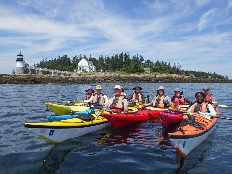 The image shows a group of people kayaking on a sunny day. They are paddling in a line, with colorful kayaks. In the background, there is a small island with a lighthouse and some trees. The water is calm and blue, and the sky is partly cloudy. The kayakers appear to be enjoying their time on the water.